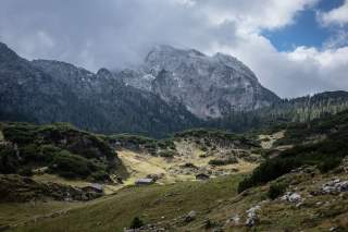 Berglandschaft mit drei kleinen Holzchalets im Vordergrund, dahinter bewaldete Hänge und ein teils wolkenverhangener, felsiger Berggipfel.