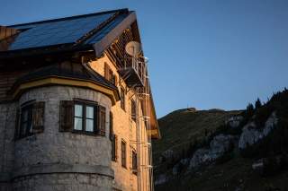 Teilansicht eines steinernen Alpenhauses mit Solarpanelen auf dem Dach im Abendlicht, im Hintergrund ein Berg mit einem Gipfelkreuz unter klarem Himmel.