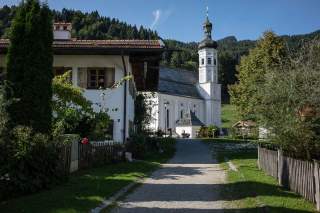 Ein schmaler Weg führt zu einer weißen Kirche mit auffälligem Turm, umgeben von einem Haus, Bäumen und bewaldeten Hügeln im Hintergrund.