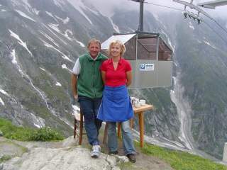 Ein Mann und eine Frau stehen lächelnd vor einer Berglandschaft mit Schneeflecken, im Hintergrund hängt eine Stahlgondel an einer Seilbahn; neben ihnen steht ein kleiner Tisch mit mehreren Tassen.