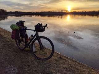 Ein mit Gepäcktaschen beladenes Fahrrad steht am Ufer eines ruhigen Gewässers bei Sonnenuntergang, während zwei Enten auf dem Wasser schwimmen.