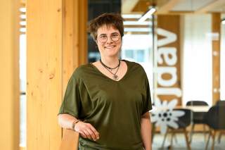 Lächelnde Person mit Brille und olivgrünem Shirt vor einer Glaswand mit dem JDAV-Logo in einem hellen Holz-Büro
