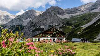 Alpenhütte mit bunten Fahnen vor hohen, teils bewachsenen Felsenbergen und blühenden Alpenrosen im Vordergrund.
