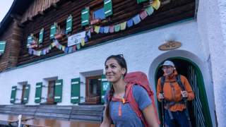 Zwei Wanderer mit Rucksäcken verlassen eine Berghütte mit grünen Fensternläden, über dem bunte Wimpelketten hängen.