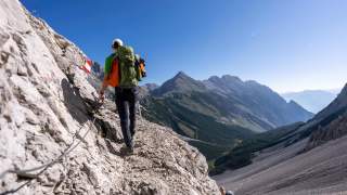 Zwei Wanderer mit Rucksäcken steigen entlang eines schroffen Bergpfads, umgeben von steilen Felsen und Bergen unter klarem blauen Himmel.