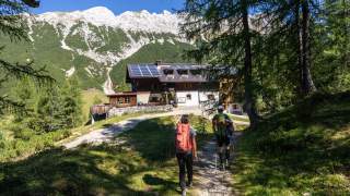 Zwei Wanderer mit Rucksäcken gehen auf einem Waldweg zu einer Berghütte mit Solarpaneelen auf dem Dach, im Hintergrund sind Berge und blauer Himmel zu sehen.