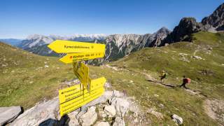 Gelber Wegweiser mit mehreren Richtungen auf einem Bergwanderweg im Karwendelgebirge, im Hintergrund zwei Wanderer auf einem Pfad und Bergpanorama.