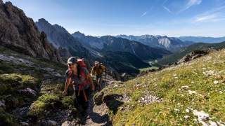 Zwei Wanderer mit Rucksäcken steigen auf einem schmalen Pfad in einer bergigen Landschaft bei klarem Himmel auf. Im Hintergrund sind zerklüftete Berge und ein Tal mit grüner Vegetation zu sehen.