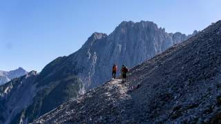 Zwei Wanderer mit Rucksäcken steigen auf einem schmalen, steinigen Pfad in den Bergen bei klarem Himmel auf. Im Hintergrund erhebt sich ein markanter, steiler Felsen.