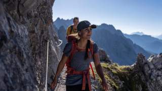 Zwei Wanderer mit Rucksäcken und Kappe gehen einen schmalen Bergpfad entlang, der von einem Felsvorsprung und Seilsicherungen begleitet wird, mit bergiger Landschaft im Hintergrund.