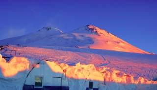 Schneebedecktes Bergmassiv im warmen Licht der untergehenden Sonne mit einem schneebedeckten Gebäude im Vordergrund. Der Himmel ist klar und tiefblau.
