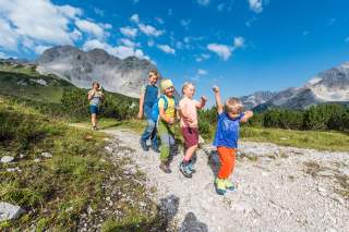 Eine Gruppe von Kindern wandert fröhlich auf einem Bergweg, im Hintergrund sind Berge und ein blauer Himmel zu sehen. Eine erwachsene Begleiterin läuft hinter ihnen her.