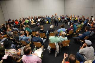 Eine große Gruppe von Menschen sitzt in einem Kreis in einem Konferenzraum, während eine Person in der Mitte mit einem Mikrofon spricht. Die Teilnehmer wirken aufmerksam und beteiligen sich am Gespräch.