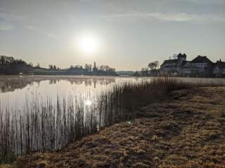 Sonnenaufgang über einem ruhigen See mit Schilf am Ufer und einer Kirche sowie einem Schloss im Hintergrund.