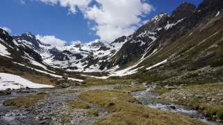 Berglandschaft mit schneebedeckten Gipfeln, einem gewölbten Tal und einem kleinen Bach, der sich durch das grüne und teils felsige Gelände schlängelt, unter einem blauen Himmel mit vereinzelten Wolken.