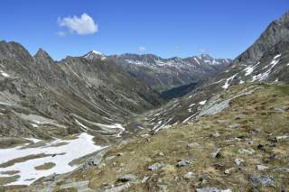 Berglandschaft mit schneebedeckten Gipfeln und einem Tal, das von Felsen und Grasflächen umgeben ist, unter blauem Himmel mit einzelnen Wolken.