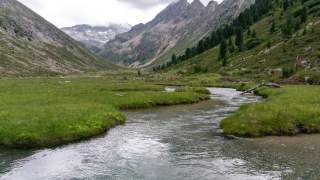 Berglandschaft mit einem klaren, sich schlängelnden Fluss, umgeben von grünen Wiesen und bewaldeten Hängen unter einem bewölkten Himmel.