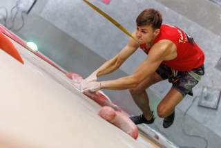 Junger Mann klettert mit rotem Trikot und schwarzer Hose an einer Indoor-Kletterwand und hält sich an roten Griffen fest.