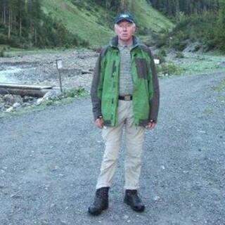 Mann in Outdoor-Bekleidung mit grüner Jacke, beige Hose und schwarzer Mütze steht auf einem felsigen Weg in einer bewaldeten Gebirgslandschaft.