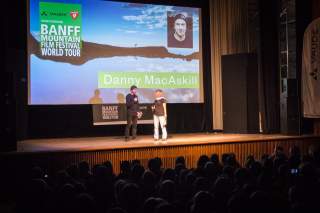 Zwei Personen stehen mit Mikrofonen auf einer Bühne vor einem großen Bildschirm mit der Aufschrift „BANFF Mountain Film Festival World Tour“ und „Danny MacAskill“, während ein Publikum zuschaut.