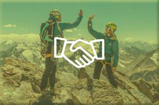 Zwei Bergsteiger mit Helmen geben sich auf einem felsigen Gipfel in alpiner Landschaft einen high five.