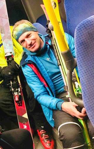 Mann in blauer Skijacke und Stirnband sitzt in einem Bus und hält Skier, neben ihm steht eine weitere Person mit Skiern und Skischuhen.