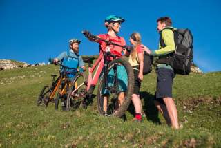 Vier Personen mit Mountainbikes und Wanderausrüstung stehen auf einer grünen Wiese unter blauem Himmel und unterhalten sich lächelnd.