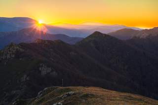 Sonnenaufgang über bewaldeten Bergen mit leuchtend orangefarbenem Himmel und sanften Hügeln im Vordergrund.