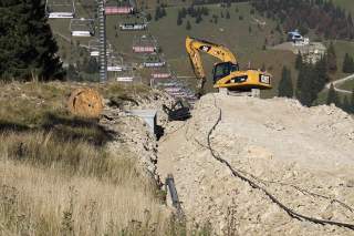 Bauarbeiten mit einem Bagger in einer aufgerissenen Erdaufschüttung neben einer Seilbahn im Gebirge.
