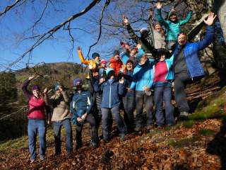 Eine Gruppe von 16 Menschen steht im Wald auf herbstlichem Laub, lacht und winkt in die Kamera, während die Sonne scheint. Im Hintergrund sind kahle Bäume und ein bewaldeter Hügel unter blauem Himmel zu sehen.