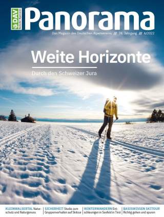 Person in gelber Jacke beim Skilanglauf auf schneebedecktem Feld mit langer Schattenbildung bei tiefstehender Sonne, im Hintergrund teilweise bewölkter Himmel und Berge.