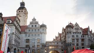 Menschen klettern an einer mobilen Kletterwand vor einem historischen Gebäude mit Türmen und einer Durchfahrt, darunter stehen Zelte und Fahnen, die auf ein Event hinweisen.