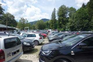 Voll besetzter Parkplatz mit vielen geparkten Autos, umgeben von Bäumen und Bergen im Hintergrund unter einem bewölkten Himmel.