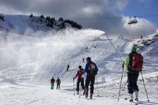 Skifahrer wandern in einer verschneiten Berglandschaft, während eine Schneekanone Schnee auf die Piste sprüht und ein Sessellift darüber fährt.