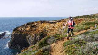 Eine Wanderin mit Rucksack und Wanderstöcken geht auf einem Pfad entlang einer felsigen Küste mit Blick auf das Meer.