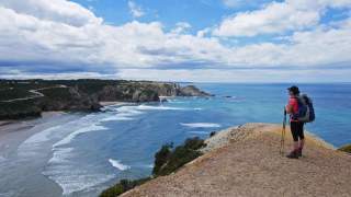 Eine Wanderin mit Rucksack und Trekkingstöcken steht auf einem Küstenpfad und blickt auf das Meer und die felsige Küstenlandschaft unter einem teils bewölkten Himmel.