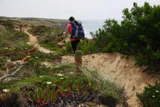 Eine Wanderin mit großem Rucksack und Trekkingstöcken läuft auf einem schmalen Pfad durch eine hügelige Küstenlandschaft mit Sanddünen und Vegetation. Im Hintergrund ist das Meer zu sehen.