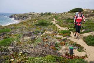 Frau mit Trekkingausrüstung wandert auf einem sandigen Küstenweg, umgeben von grüner Vegetation und mit Blick auf das Meer.