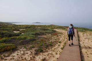 Eine Frau mit Rucksack und Wanderstöcken geht auf einem Holzsteg durch eine karge Landschaft mit niedriger Vegetation an der Küste entlang, im Hintergrund Meer und Inseln.