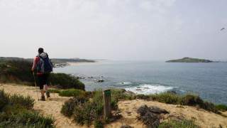 Eine Person mit Rucksack und Wanderstöcken läuft auf einem sandigen Pfad entlang einer Küste mit Blick auf das Meer und eine kleine Insel. Im Hintergrund ist ein bewölkter Himmel zu sehen.