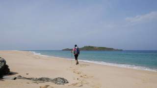 Eine allein wandernde Person mit Rucksack und Wanderstock geht am Strand entlang, vor ihr das Meer mit einer kleinen Insel im Hintergrund.