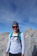Junge Frau mit Sonnenbrille, Stirnband und Rucksack steht lächelnd vor einer felsigen Berglandschaft unter blauem Himmel.