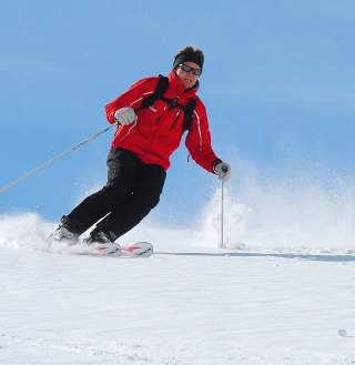 Person in roter Jacke und schwarzer Hose fährt mit Skiern im Schnee, vor blauem Himmel.