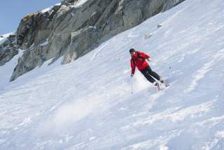 Eine Person in roter Skijacke fährt mit Skiern einen schneebedeckten Hang neben einer felsigen Bergwand hinunter.