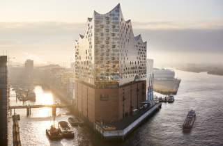 Die Elbphilharmonie in Hamburg steht auf einem ehemaligen Kaispeicher, umgeben von Wasser und Schiffen bei Sonnenuntergang. Das moderne Gebäude vereint eine gläserne Oberstruktur mit einer historischen Backsteinbasis.