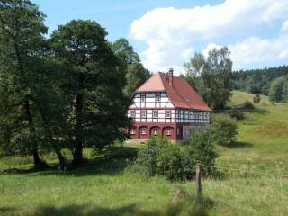 Fachwerkhaus mit rotem Dach in einer grünen, hügeligen Landschaft umgeben von Bäumen und Wiesen unter blauem Himmel mit Wolken.