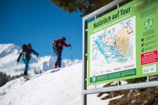 Informationsschild mit Karte und Hinweisen für naturverträgliche Skitouren und Schneeschuhgehen in den Alpen, im Hintergrund zwei Personen beim Aufstieg im Schnee vor Bergkulisse.