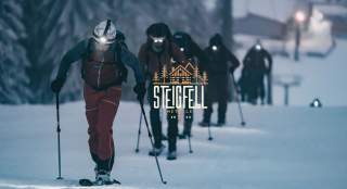 Mehrere Personen in Winterkleidung und Stirnlampen wandern bei Dunkelheit mit Schneeschuhen durch eine verschneite Landschaft.