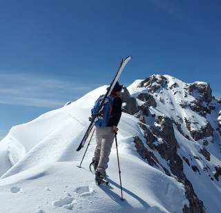 Eine Person mit Skiern auf dem Rücken und Wanderstöcken steigt auf einem schneebedeckten Berggipfel bei blauem Himmel auf.