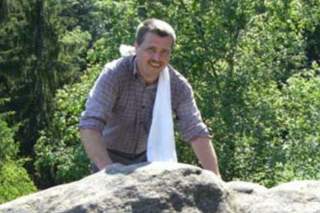 Ein Mann mit Schnurrbart und kariertem Hemd steht lächelnd hinter einem großen Felsen im Freien, mit einem weißen Tuch über der Schulter und grüner Waldlandschaft im Hintergrund.